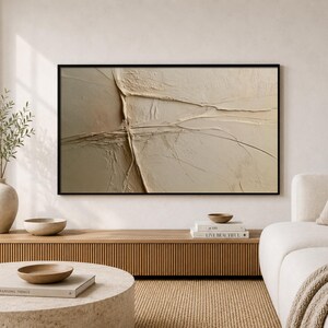May include: A rectangular wall art piece featuring a textured, beige, and cream-colored abstract composition with raised, linear ridges and deep grooves, framed in a thin black border, displayed horizontally on a light-colored wall.