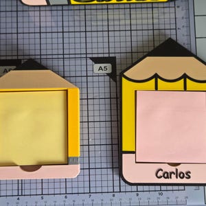 Puede incluir: Dos soportes para notas adhesivas con forma de lápiz hechos de plástico amarillo, negro y rosa. El soporte izquierdo contiene una nota amarilla, mientras que el derecho contiene una nota rosa y lleva el nombre Carlos impreso en letras negras.