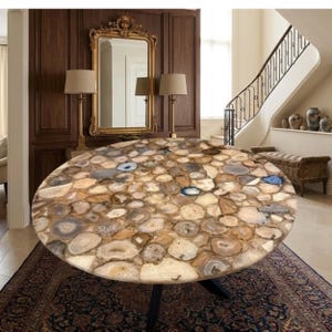 Op de afbeelding: Een ronde tafel met een mozaïekachtig oppervlak van gepolijste agaatstenen in bruin, beige en blauw. De tafel heeft een zwart metalen onderstel en staat op een patroonkleed. De tafel heeft een diameter van ongeveer 122 cm.