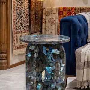 May include: A round side table constructed from polished labradorite stone, featuring a mosaic of dark gray and iridescent blue segments. The table has a cylindrical base and a flat top, with the text Stoniracraftsindia visible on the surface.