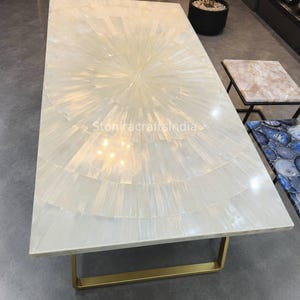 May include: A rectangular dining table featuring a white selenite stone top with a sunburst pattern of inlaid segments. The table is supported by a gold-toned metal base. The text StoniracraftsIndia is visible on the surface of the table.