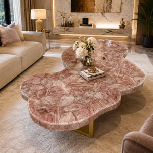 May include: A coffee table featuring a large, irregular, cloud-shaped top made of polished pink rose quartz stone. The surface displays a natural crystalline texture with varied shades of pink and white. It is supported by a gold-toned metal base.