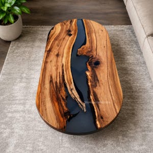 Puede incluir: Una mesa de centro ovalada con dos tablones de madera de borde natural separados por una incrustación de resina gris oscuro lisa. La madera muestra patrones de veta y nudos prominentes. El texto StoniracraftsIndia es visible en la resina.