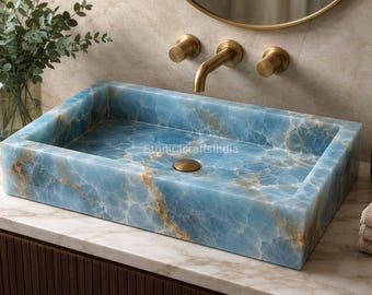 Può includere: Un lavabo rettangolare realizzato in pietra onice blu con venature naturali bianche e dorate. La vasca presenta un tappo di scarico circolare centrale con finitura oro spazzolato, abbinato alla rubinetteria a parete sopra il lavabo.