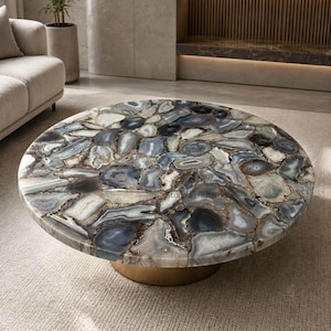 Op de afbeelding: Een ronde salontafel met een tafelblad dat bestaat uit talloze gepolijste, meerkleurige grijze, witte en zwarte agaatstenen plakken in een mozaïekpatroon. De tafel rust op een massieve, cilindervormige goudkleurige metalen voet.