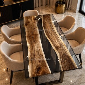 Op de afbeelding: Een rechthoekige eettafel met twee houten bladen met natuurlijke randen, gescheiden door een centrale strook van donkere, doorschijnende hars. De tafel heeft een zwart metalen frame en zes beige gestoffeerde stoelen. Tekst: Stoniracraftsindia.