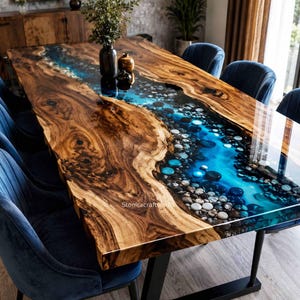 Könnte beinhalten: Ein rechteckiger Esstisch mit einer Massivholzplatte mit Naturkante und einem zentralen Flussdesign aus blauem Harz und glatten Flusskieselsteinen. Der Tisch steht auf schwarzen Metallbeinen und ist von sechs blauen Samtstühlen umgeben.