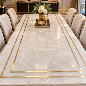 Puede incluir: Una mesa de comedor rectangular con una superficie de mármol blanco pulido con bordes incrustados de metal dorado y una base de metal dorado. La mesa está rodeada de sillas tapizadas y muestra una bandeja central con flores y velas. El texto dice StoniracraftsIndia.