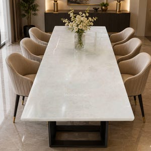 Puede incluir: Una mesa de comedor rectangular con una superficie de piedra blanca pulida y una base de metal negro, rodeada por seis sillas tapizadas en color beige con costuras verticales y patas negras. Un florero de cristal con flores blancas.