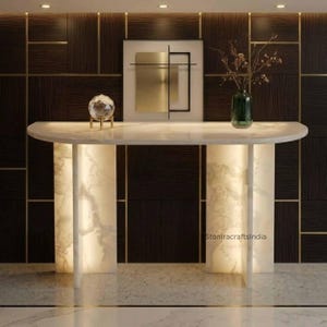 May include: A semi-circular console table made of white marble with integrated internal lighting in the base. On the surface sits a clear glass sphere on a gold stand and a green glass vase with dried branches. Text reads StoniracraftsIndia.