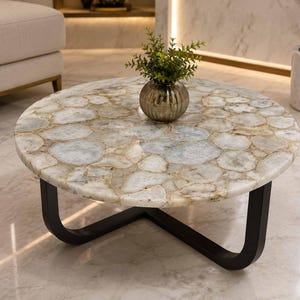 Op de afbeelding: Een ronde salontafel met een tafelblad samengesteld uit ingelegde doorschijnende witte en beige agaatplakken. De tafel wordt ondersteund door een zwart metalen onderstel met een gebogen, open frameontwerp. Een kleine groene plant staat in een geribbelde vaas.
