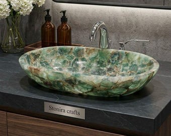 Può includere: Un lavabo da bagno ovale realizzato in pietra naturale verde e marrone con finitura lucida. Il lavabo presenta una marcata consistenza cristallina multicolore ed è montato su un piano scuro con una targhetta metallica con la scritta Stonira crafts.