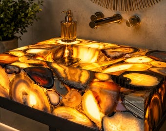 Può includere: Un lavabo rettangolare costruito con fette di pietra agata retroilluminate e multicolori nelle tonalità ambra, oro e marrone. Il lavabo presenta una finitura lucida ed è montato su una struttura in metallo nero, accompagnato da un rubinetto dorato.