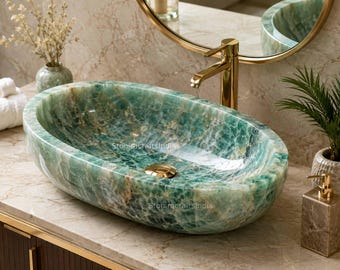 Può includere: Un lavabo ovale realizzato in pietra verde lucidata con venature naturali e finitura brillante. Il bacino presenta una piletta di scarico in metallo dorato centrata sul fondo. Il testo StoniracraftsIndia è visibile sulla superficie interna.