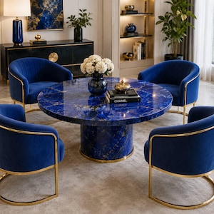 Op de afbeelding: Een ronde eettafel met een blauw marmeren blad en een cilindrische voet, omringd door vier blauwe fluwelen stoelen met gouden metalen frames. Een donkerblauwe vaas met witte bloemen en een zwart boek met de titel CHANEL op tafel.