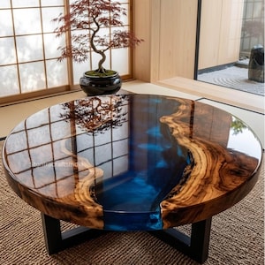 May include: A round coffee table featuring a natural wood slab with a polished finish and a central river-like inlay of translucent blue resin. The table is supported by a black metal base and displays the text StonecraftsIndia on the surface.