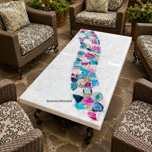 May include: A rectangular coffee table with a white quartz surface featuring a central, curved inlay of multicoloured agate slices in shades of pink, blue, and teal. The table has a dark metal base and includes the text StoniracraftsIndia.