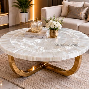 Op de afbeelding: Een ronde salontafel met een oppervlak van wit parelmoer ingelegd in een radiaal patroon, ondersteund door een gepolijst goudkleurig metalen onderstel met een gebogen kruisontwerp. Het tafeloppervlak toont de tekst StoniracraftsIndia.