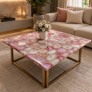 May include: A square coffee table featuring a tabletop composed of polished pink and white agate slices with gold-toned metal legs. A white vase filled with white flowers sits in the center of the table. The text Stoniracraftstudia is visible.
