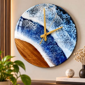 May include: A circular wall clock featuring a natural wood base on the bottom half and a glossy resin ocean design on the top half. The resin depicts deep blue water with white foam waves. It has three thin, gold-colored metal clock hands.