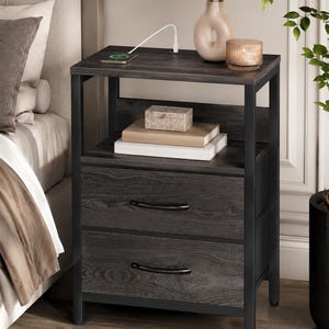 May include: A dark gray nightstand with two drawers and a top shelf. The nightstand has a black metal frame and curved drawer pulls. A phone is charging on the top shelf, along with a vase, candle, and decorative box.