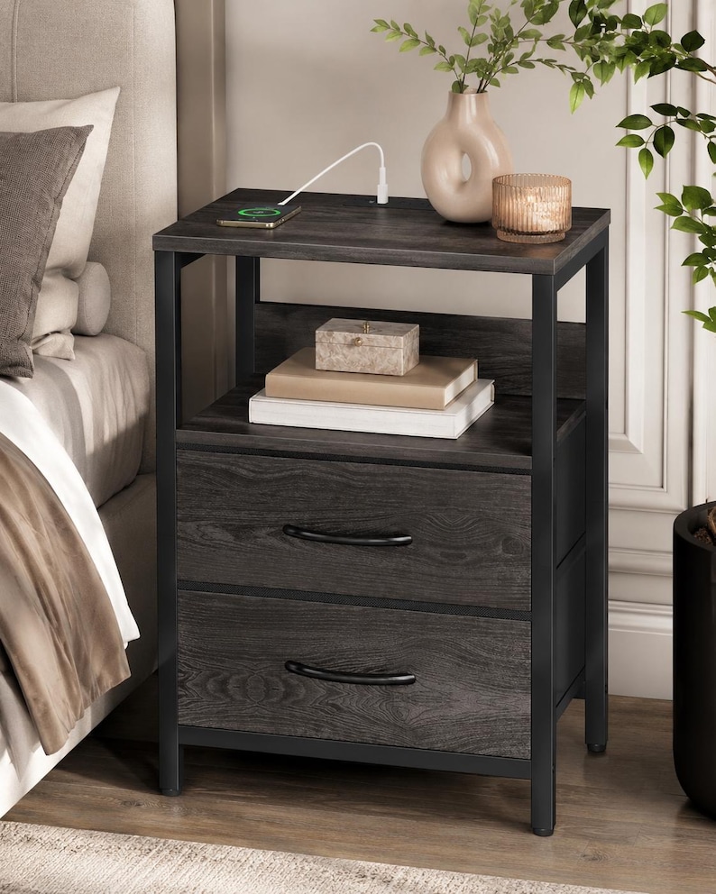 May include: A dark gray nightstand with two drawers and a top shelf. The nightstand has a black metal frame and curved drawer pulls. A phone is charging on the top shelf, along with a vase, candle, and decorative box.