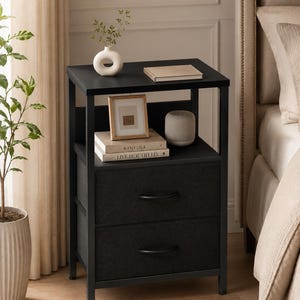 May include: A black nightstand with two drawers and a shelf. The top shelf holds a white vase with greenery, a book, and a small framed picture. The bottom shelf has a stack of books and a gray cylindrical vase. The nightstand is next to a bed.