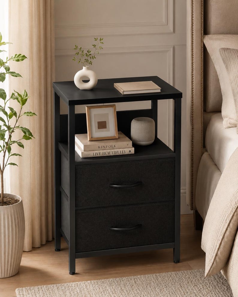 May include: A black nightstand with two drawers and a shelf. The top shelf holds a white vase with greenery, a book, and a small framed picture. The bottom shelf has a stack of books and a gray cylindrical vase. The nightstand is next to a bed.