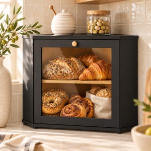 May include: A black corner bread box with a clear glass front door and a light wood interior. The two-tier storage unit holds a variety of baked goods including a large seeded loaf, croissants, bagels, and cinnamon rolls inside a fabric liner.