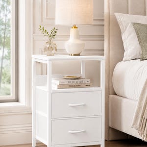 May include: A white nightstand with two drawers and a shelf, topped with a lamp and a small vase with greenery. The lamp has a beige shade and a white base with gold accents. Books are on the shelf. The nightstand is next to a bed.