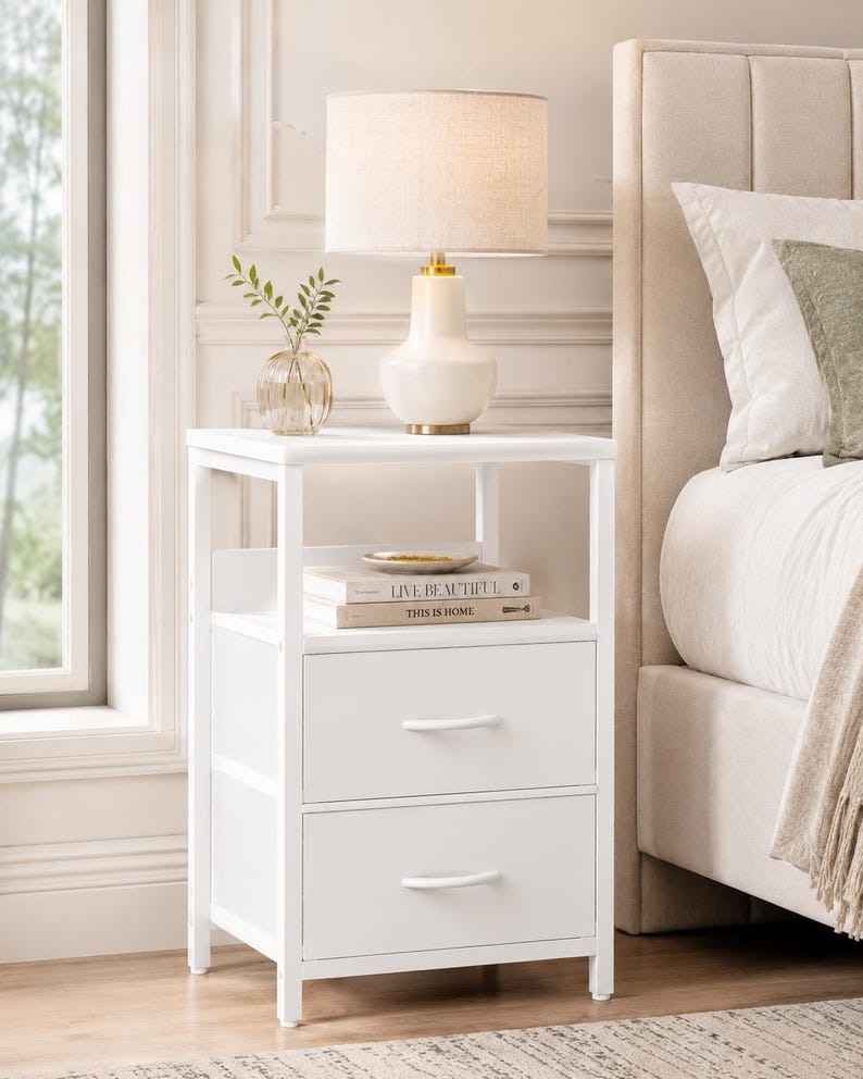 May include: A white nightstand with two drawers and a shelf, topped with a lamp and a small vase with greenery. The lamp has a beige shade and a white base with gold accents. Books are on the shelf. The nightstand is next to a bed.