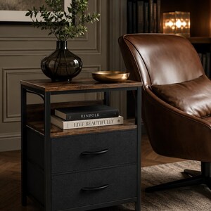 May include: A dark brown side table with a black metal frame. The table has a wooden top and a shelf with two books, "KINFOLK" and "LIVE BEAUTIFULLY". Below the shelf are two drawers with black fabric fronts. A dark vase with greenery and a gold bowl sit on top.