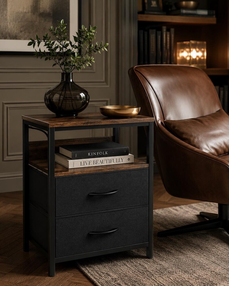 May include: A dark brown side table with a black metal frame. The table has a wooden top and a shelf with two books, "KINFOLK" and "LIVE BEAUTIFULLY". Below the shelf are two drawers with black fabric fronts. A dark vase with greenery and a gold bowl sit on top.