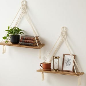 May include: Two rectangular wooden floating shelves suspended by cream-colored macrame rope with circular wooden rings at the top. The top shelf holds a green plant in a black pot and three stacked books. The bottom shelf holds a brown mug, a gold hourglass, and a framed botanical print.