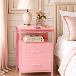 May include: A pink nightstand with two drawers, a shelf, and a top surface. A white lamp with a pleated shade sits on top, along with a vase of pink flowers, a phone, and books. The nightstand is next to a bed.