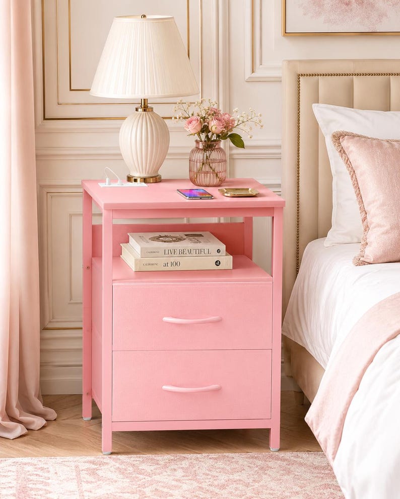 May include: A pink nightstand with two drawers, a shelf, and a top surface. A white lamp with a pleated shade sits on top, along with a vase of pink flowers, a phone, and books. The nightstand is next to a bed.