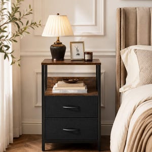May include: A two-drawer nightstand with a dark wood top and black metal frame. A lamp with a pleated shade sits on top, along with a framed picture and a candle. The drawers are black with curved handles. A bed with a beige headboard is in the background.