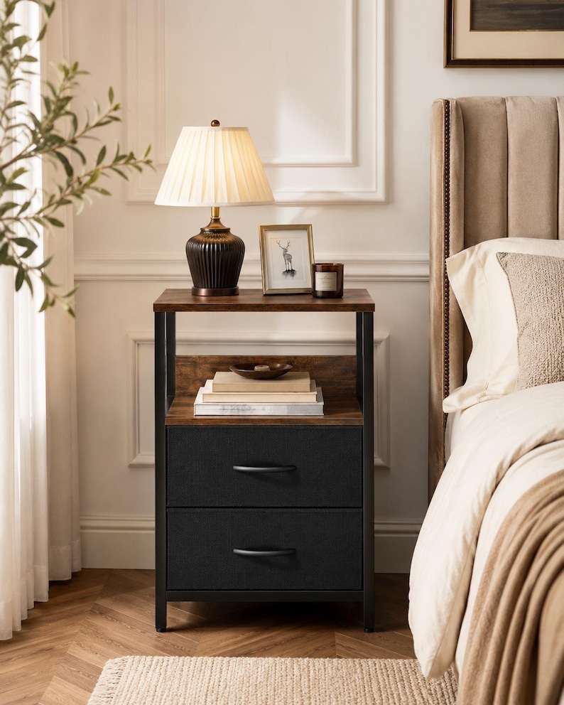May include: A two-drawer nightstand with a dark wood top and black metal frame. A lamp with a pleated shade sits on top, along with a framed picture and a candle. The drawers are black with curved handles. A bed with a beige headboard is in the background.