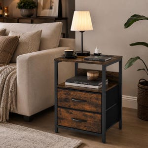 May include: A rustic-style side table with two drawers, a black metal frame, and a distressed wood finish. A small lamp with a pleated shade sits on top, along with a coffee cup and books. The table is 24 inches tall.