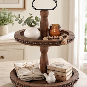 May include: A two-tiered, wooden serving tray with a dark finish. The top tier holds a small white vase with greenery, a lit candle in a glass jar, and wooden beads. The bottom tier features folded linen napkins, a ceramic bird, and small books.