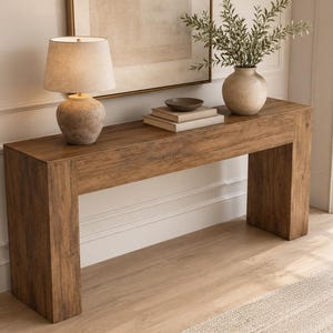 May include: A rectangular console table made of brown wood with a visible grain pattern. The table features a thick, block-style frame with two wide legs. On top sits a table lamp with a textured base, two stacked books, a small bowl, and a vase.