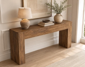 May include: A rectangular wooden console table with a natural brown wood grain finish. The table features a thick, block-style frame with four legs. On top sits a beige lamp, two stacked books, a small dark bowl, and a vase with green foliage.