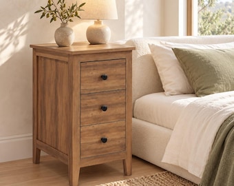 May include: A wooden nightstand featuring three drawers with small black round knobs. The nightstand has a light brown wood grain finish and stands on four legs. A ceramic vase with olive branches and a table lamp sit on the top surface.
