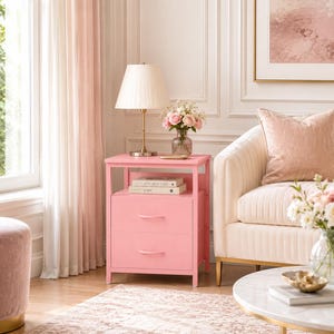 May include: A pink bedside table with two drawers and a shelf. A lamp with a cream shade sits on top, along with books and a vase of pink roses. The table is next to a cream-colored sofa and a round coffee table.