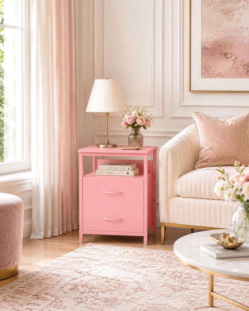 May include: A pink bedside table with two drawers and a shelf. A lamp with a cream shade sits on top, along with books and a vase of pink roses. The table is next to a cream-colored sofa and a round coffee table.