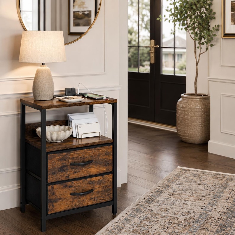 May include: A two-drawer nightstand with a brown, wood-grain finish and black metal frame. A beige lamp sits on top, along with a charging station and a smartphone. A decorative bowl and a stack of white cards are on the middle shelf.