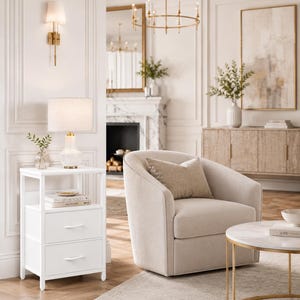 May include: A bright, airy living room with neutral tones. Features include a white nightstand with two drawers, a cream-colored armchair, a marble-topped coffee table, and a gold chandelier. A lamp with a linen shade sits on the nightstand.