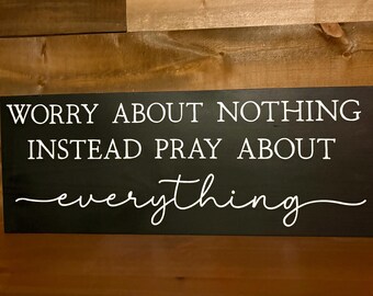 May include: A rectangular black wooden sign with white lettering. The text reads "WORRY ABOUT NOTHING INSTEAD PRAY ABOUT everything" in a mix of block and cursive fonts. The sign is displayed against a brown wooden background.