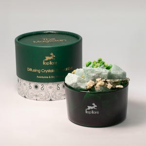 May include: A dark green container with a lid, next to a smaller dark green container filled with pale green crystals and dried flowers. The larger container has text that reads "The Magician" and "Hop Hare". The smaller container also has the "Hop Hare" logo.