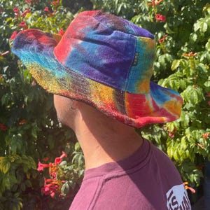 May include: A bucket hat featuring a vibrant tie-dye pattern in shades of red, blue, purple, yellow, and orange. The hat has a wide, downward-sloping brim and a small black fabric tag on the side with the word HEMP printed in white letters.